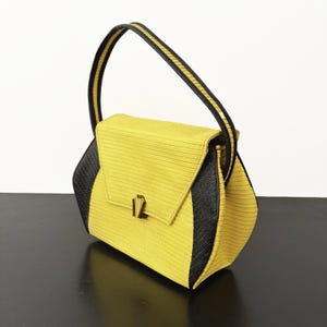 Op de afbeelding: Een gele en zwarte handtas met een gestructureerd ontwerp. De tas heeft een getextureerde gele body met zwarte zijpanelen en een zwarte handgreep met een gouden streep. Het heeft een rechthoekige flap met een gouden sluiting.