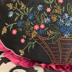K&ouml;nnte beinhalten: Ein schwarzes Kissen mit rosa R&uuml;schenbesatz zeigt gestickte Blumen in einem Korb. Die Blumen sind in Blaut&ouml;nen, Rosa und Gelb gehalten.