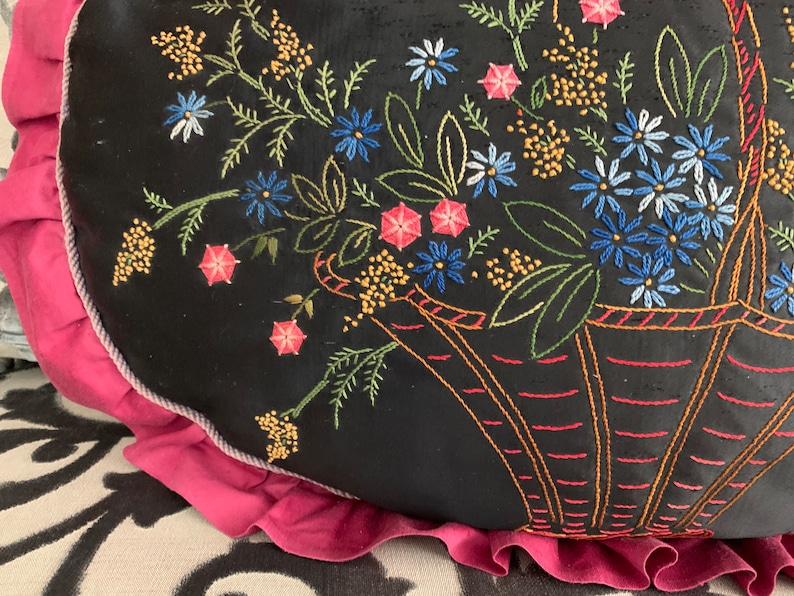 K&ouml;nnte beinhalten: Ein schwarzes Kissen mit rosa R&uuml;schenbesatz zeigt gestickte Blumen in einem Korb. Die Blumen sind in Blaut&ouml;nen, Rosa und Gelb gehalten.