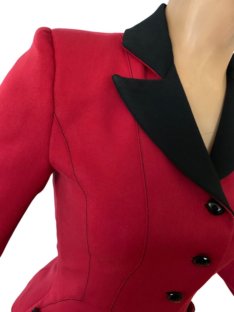 Puede incluir: Una chaqueta roja con cuello negro y botones negros. La chaqueta tiene una silueta ajustada y un cierre de doble botonadura.