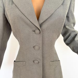Puede incluir: Un blazer gris claro con un corte entallado. El blazer presenta una solapa con muescas, tres botones a juego y dos bolsillos delanteros. La tela tiene un aspecto sutilmente texturizado. Las mangas son largas y el blazer se lleva sobre una prenda roja.