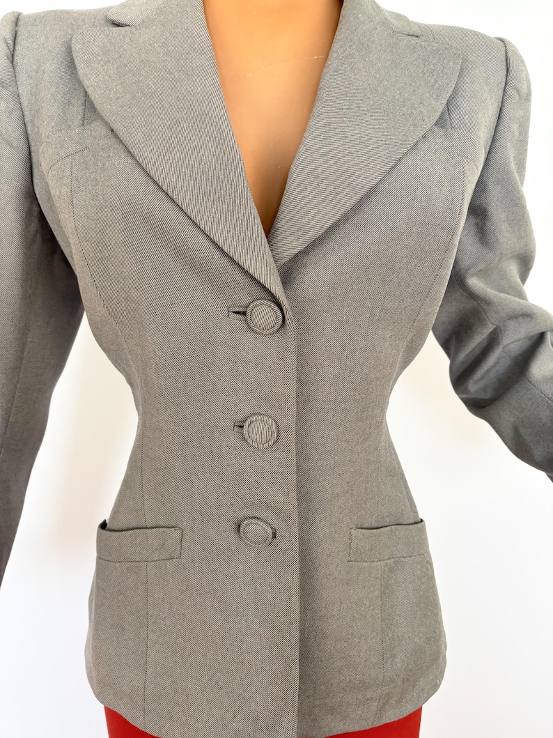 Puede incluir: Un blazer gris claro con un corte entallado. El blazer presenta una solapa con muescas, tres botones a juego y dos bolsillos delanteros. La tela tiene un aspecto sutilmente texturizado. Las mangas son largas y el blazer se lleva sobre una prenda roja.