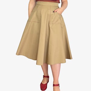 Op de afbeelding: Een beige knielange rok met grote voorzakken en contrasterende stiksels. De rok heeft een uitlopende A-lijn vorm. De persoon draagt rode sandalen. De top is gestreept en off-shoulder.
