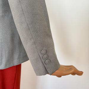 Puede incluir: Manga de blazer gris con tres botones a juego. La manga est&aacute; en &aacute;ngulo, revelando un vistazo de tela roja debajo. El blazer parece estar hecho de un material texturizado.