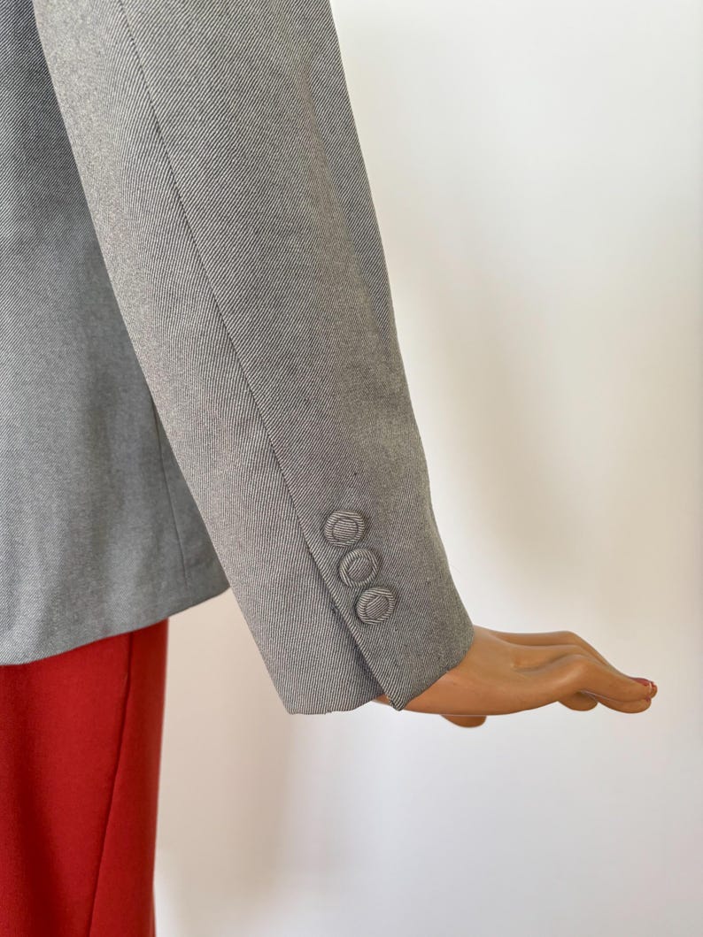 Puede incluir: Manga de blazer gris con tres botones a juego. La manga est&aacute; en &aacute;ngulo, revelando un vistazo de tela roja debajo. El blazer parece estar hecho de un material texturizado.