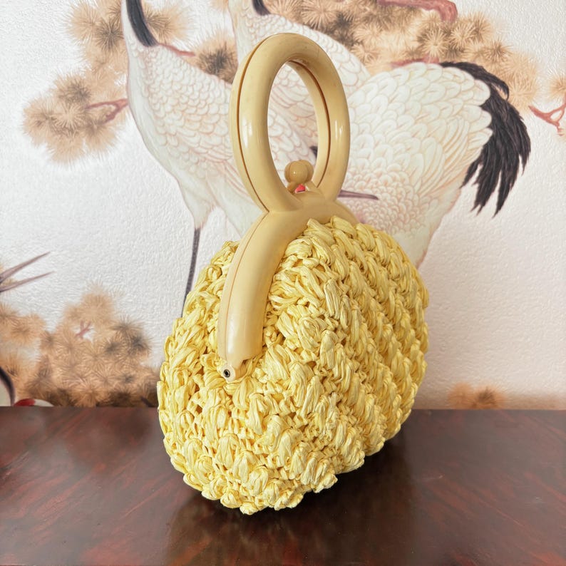 May include: A light yellow woven handbag with a rounded shape and a cream-colored handle. The bag has a textured, layered design. The handle is arched and features a small, round accent. The background features a painting of white birds.