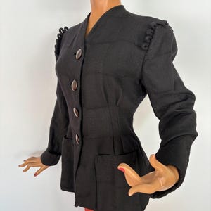 May include: A black blazer with a tailored fit, featuring a V-neckline and four brown buttons. The blazer has two front pockets and decorative shoulder detailing. The jacket is displayed on a mannequin.