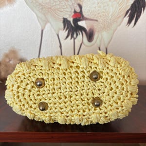 May include: A woven, pale yellow handbag with a rectangular base. The bag features four metal feet and a textured, open weave design. The bag is made of a straw-like material and appears to be handmade.