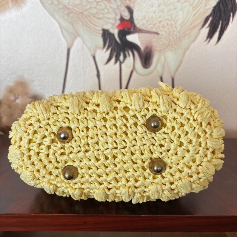 May include: A woven, pale yellow handbag with a rectangular base. The bag features four metal feet and a textured, open weave design. The bag is made of a straw-like material and appears to be handmade.