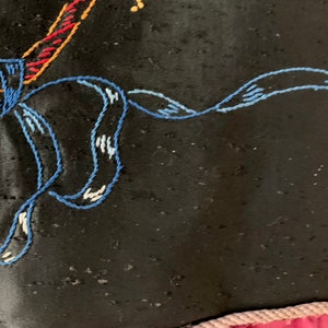 K&ouml;nnte beinhalten: Nahaufnahme eines schwarzen Stoffes mit blauen, roten und gelben Stickereien. Die Stickerei hat die Form einer Blume mit einer Schleife.