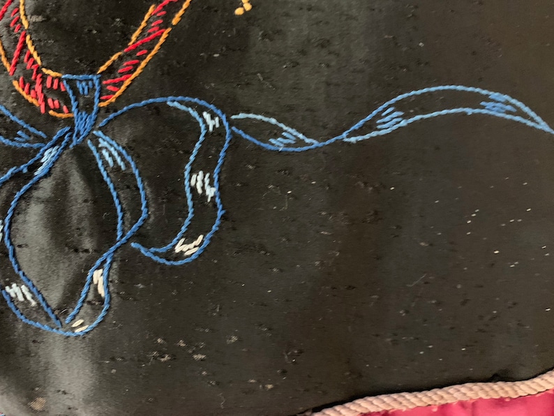 K&ouml;nnte beinhalten: Nahaufnahme eines schwarzen Stoffes mit blauen, roten und gelben Stickereien. Die Stickerei hat die Form einer Blume mit einer Schleife.