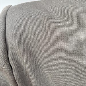 Puede incluir: Primer plano de una blazer gris, que muestra el hombro y la parte superior de la manga. La tela tiene un sutil patr&oacute;n de tejido diagonal. La blazer parece estar hecha de un material texturizado.