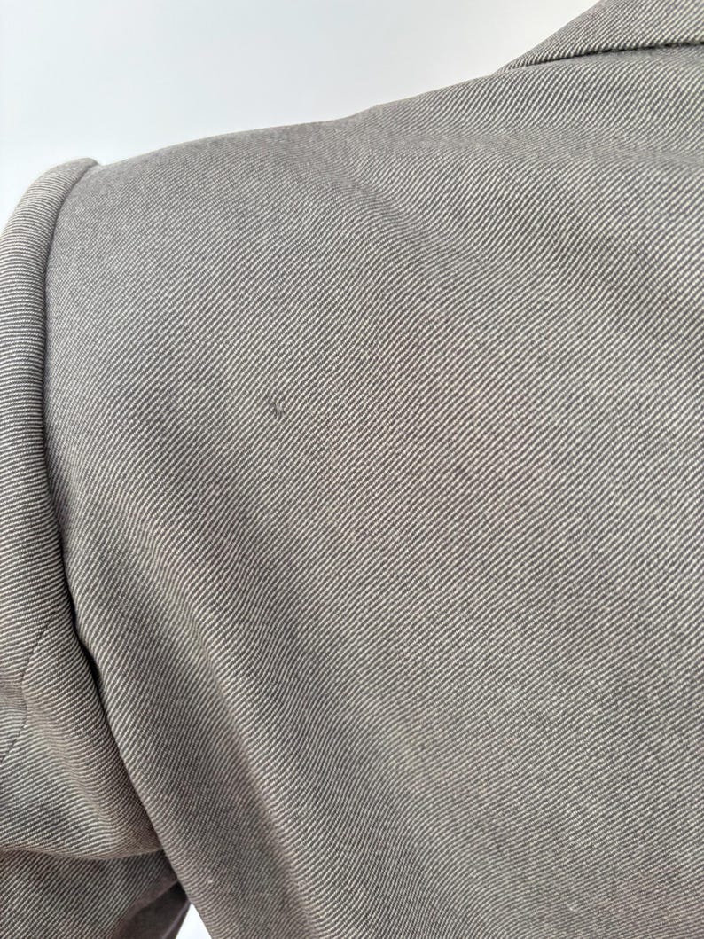 Puede incluir: Primer plano de una blazer gris, que muestra el hombro y la parte superior de la manga. La tela tiene un sutil patr&oacute;n de tejido diagonal. La blazer parece estar hecha de un material texturizado.