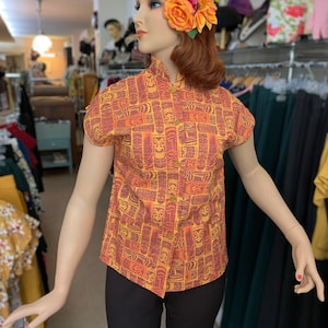 May include: A short-sleeved, orange and brown patterned shirt with a mandarin collar and button fastening. The shirt features a tiki-inspired design with images of faces and geometric patterns.