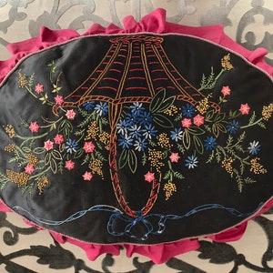 K&ouml;nnte beinhalten: Ein schwarzes Samtkissen mit einem rosa R&uuml;schenrand zeigt ein gesticktes Design einer Lampenkappe mit Blumen. Die Lampenkappe ist rot umrandet und die Blumen sind in den Farben Rosa, Blau und Gr&uuml;n gehalten.