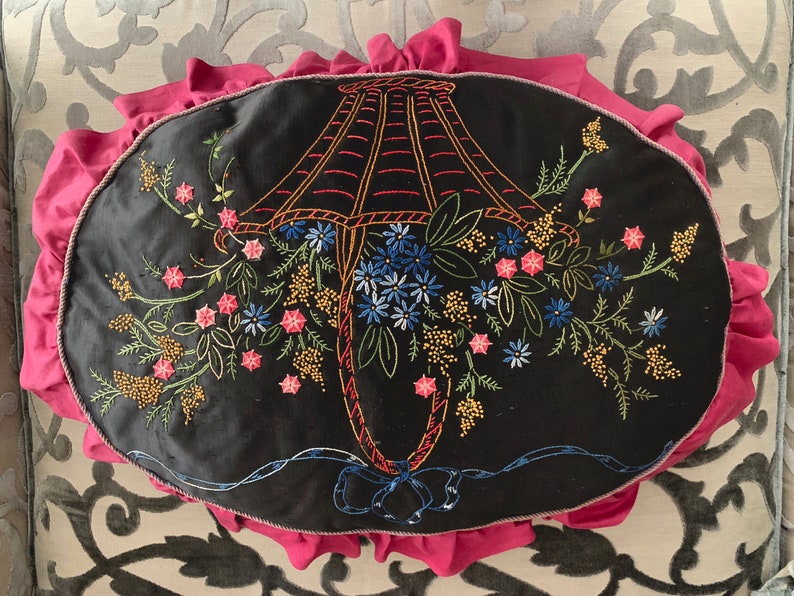 K&ouml;nnte beinhalten: Ein schwarzes Samtkissen mit einem rosa R&uuml;schenrand zeigt ein gesticktes Design einer Lampenkappe mit Blumen. Die Lampenkappe ist rot umrandet und die Blumen sind in den Farben Rosa, Blau und Gr&uuml;n gehalten.