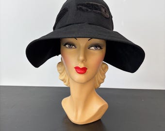 May include: A black hat with a wide brim and a rounded crown. The hat has two dark velvet accents on the crown. The hat is displayed on a mannequin head with red lipstick and blonde hair.