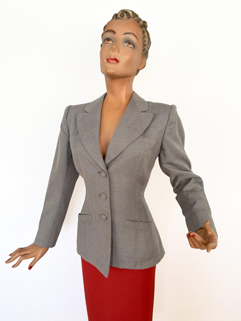 Puede incluir: Una blazer gris con un corte entallado, con cierre de dos botones y dos bolsillos delanteros. La blazer se exhibe en un maniqu&iacute; con una falda roja. El maniqu&iacute; tiene un peinado vintage y mira hacia arriba.