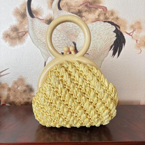 May include: A vintage, light yellow woven handbag with a cream-colored handle. The bag features a rounded shape and a clasp closure. The handle is a large, circular design. The bag is set against a backdrop of a floral design.