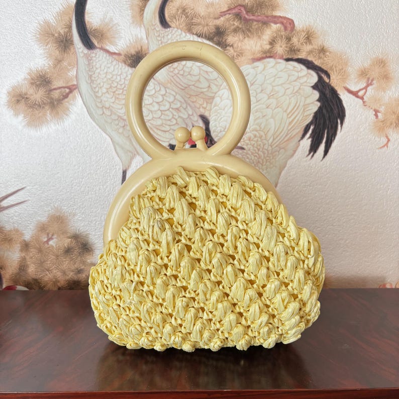 May include: A vintage, light yellow woven handbag with a cream-colored handle. The bag features a rounded shape and a clasp closure. The handle is a large, circular design. The bag is set against a backdrop of a floral design.