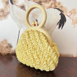 May include: A vintage, pale yellow straw handbag with a cream-colored handle and clasp. The bag has a woven texture and a rounded shape. The handle is a large, circular design. The background features a floral wallpaper.