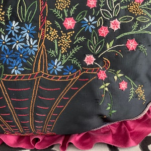 K&ouml;nnte beinhalten: Ein schwarzes Kissen mit rosa R&uuml;schenbesatz zeigt ein gesticktes Design eines Korbes, der &uuml;berquillt mit bunten Blumen. Die Blumen sind in den Farben Rosa, Blau, Gr&uuml;n und Gelb gehalten.
