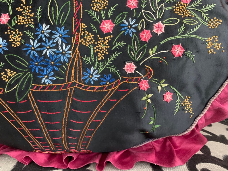K&ouml;nnte beinhalten: Ein schwarzes Kissen mit rosa R&uuml;schenbesatz zeigt ein gesticktes Design eines Korbes, der &uuml;berquillt mit bunten Blumen. Die Blumen sind in den Farben Rosa, Blau, Gr&uuml;n und Gelb gehalten.