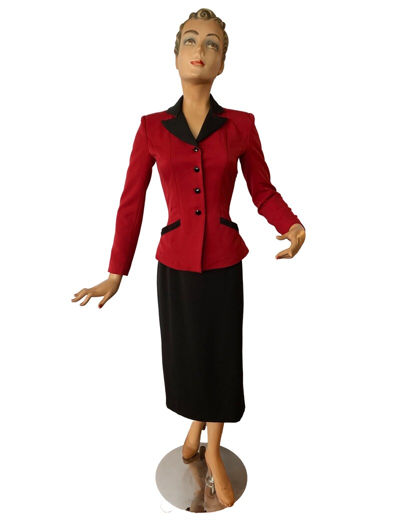 Puede incluir: Una chaqueta roja con cuello negro y botones negros, combinada con una falda negra. La chaqueta tiene una silueta ajustada y la falda es de largo hasta la rodilla.