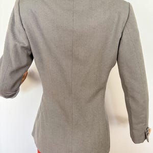 Puede incluir: Un blazer gris con un corte entallado, con solapa de muesca y mangas largas. La vista trasera muestra el dise&ntilde;o estructurado y un cierre de un solo bot&oacute;n en las mangas. El blazer se combina con una prenda roja.