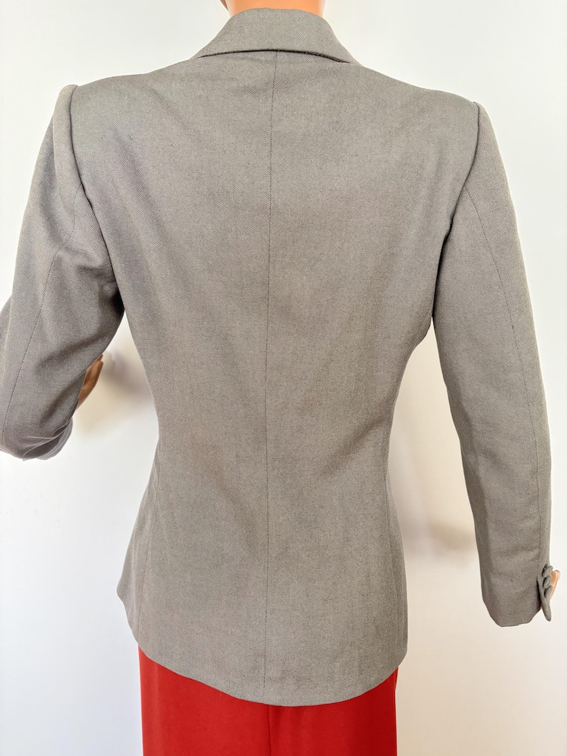Puede incluir: Un blazer gris con un corte entallado, con solapa de muesca y mangas largas. La vista trasera muestra el dise&ntilde;o estructurado y un cierre de un solo bot&oacute;n en las mangas. El blazer se combina con una prenda roja.