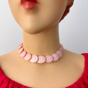 Op de afbeelding: Een roze chokerketting van plastic, met een gegolfd ontwerp. De ketting is gemaakt van een reeks overlappende cirkels.