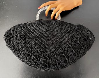Puede incluir: Bolso de mano negro de ganchillo con un asa transparente y redondeada. El bolso presenta un patrón detallado y texturizado y una forma redondeada. El asa está sujeta por una mano con esmalte de uñas rojo. El bolso está sobre una superficie negra.