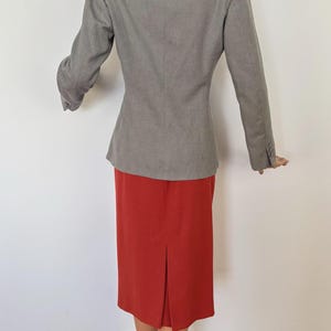 Puede incluir: Un blazer gris y una falda roja. El blazer tiene mangas largas y un corte entallado. La falda es hasta la rodilla con una abertura trasera. El maniqu&iacute; lleva tacones altos marrones.