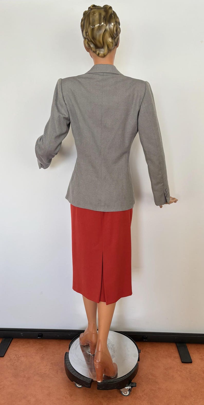 Puede incluir: Un blazer gris y una falda roja. El blazer tiene mangas largas y un corte entallado. La falda es hasta la rodilla con una abertura trasera. El maniqu&iacute; lleva tacones altos marrones.