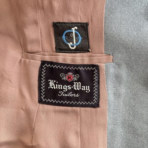 Puede incluir: Primer plano del interior de una chaqueta a medida. El forro es de color rosa apagado, con tela gris a la derecha. Dos parches bordados est&aacute;n cosidos: uno con un logotipo, el otro con el texto "Kings-Way Tailors".