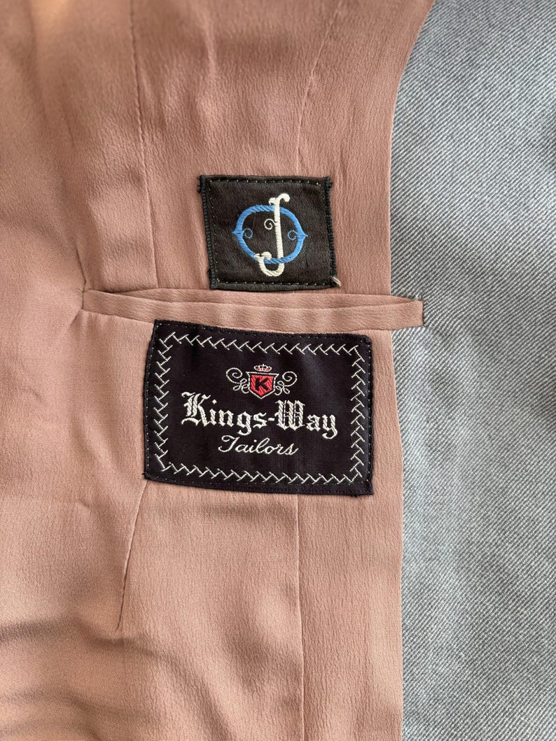 Puede incluir: Primer plano del interior de una chaqueta a medida. El forro es de color rosa apagado, con tela gris a la derecha. Dos parches bordados est&aacute;n cosidos: uno con un logotipo, el otro con el texto "Kings-Way Tailors".
