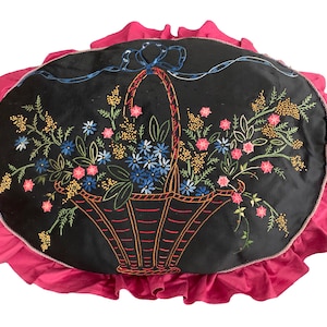 K&ouml;nnte beinhalten: Ein schwarzes Samtkissen mit einem rosa R&uuml;schenrand zeigt ein gesticktes Design eines Korbes, der mit bunten Blumen &uuml;berquillt.