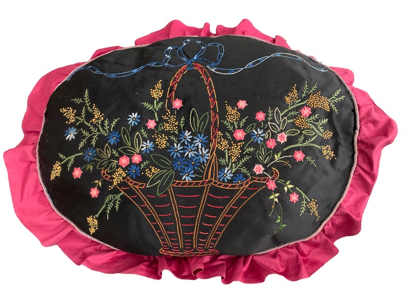 K&ouml;nnte beinhalten: Ein schwarzes Samtkissen mit einem rosa R&uuml;schenrand zeigt ein gesticktes Design eines Korbes, der mit bunten Blumen &uuml;berquillt.
