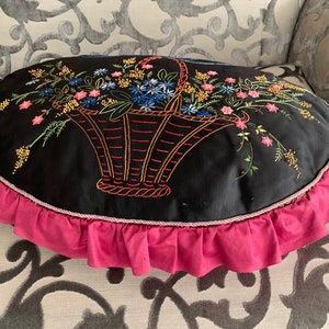 K&ouml;nnte beinhalten: Ein schwarzes ovales Kissen mit einem rosa R&uuml;schenbesatz. Das Kissen zeigt ein gesticktes Design eines Korbes, der mit bunten Blumen &uuml;berquillt.