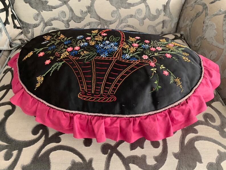 K&ouml;nnte beinhalten: Ein schwarzes ovales Kissen mit einem rosa R&uuml;schenbesatz. Das Kissen zeigt ein gesticktes Design eines Korbes, der mit bunten Blumen &uuml;berquillt.