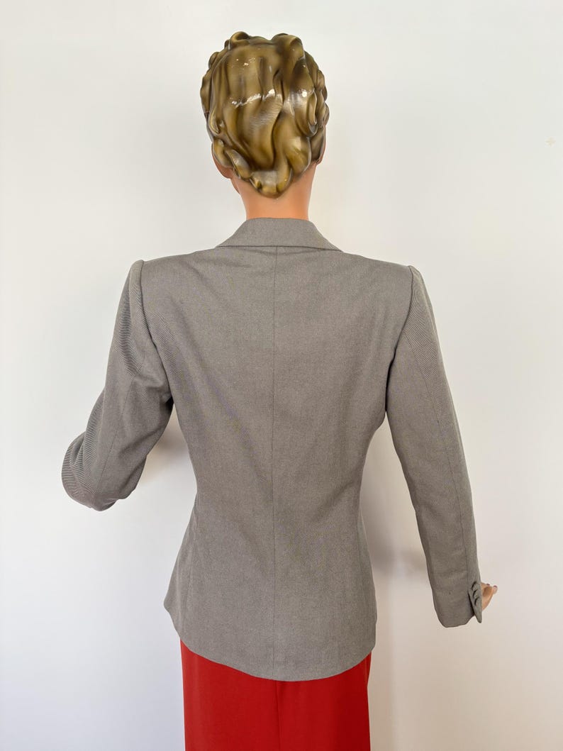 Puede incluir: Un blazer gris con un corte a medida, con mangas largas y un dise&ntilde;o cl&aacute;sico. El blazer se combina con una falda roja, creando una combinaci&oacute;n de colores contrastantes. La vista trasera muestra la estructura y el estilo de la prenda.