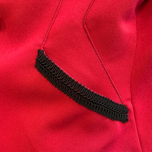 Puede incluir: Primer plano de una chaqueta de tela roja con un detalle de ribete negro. El ribete es una trenza decorativa que recorre el borde de un bolsillo.