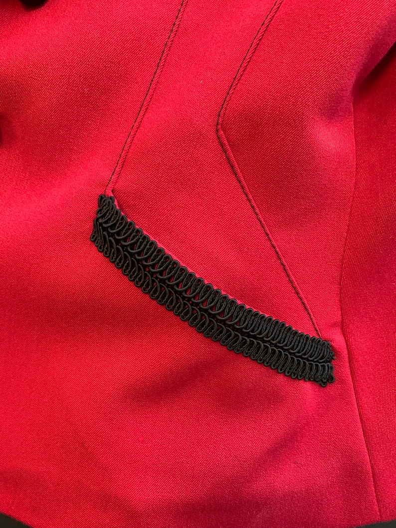 Puede incluir: Primer plano de una chaqueta de tela roja con un detalle de ribete negro. El ribete es una trenza decorativa que recorre el borde de un bolsillo.