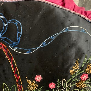 K&ouml;nnte beinhalten: Ein schwarzes Kissen mit gestickten Blumendesigns in den Farben Rosa, Gelb, Gr&uuml;n und Blau. Ein blaues Band ist oben auf dem Kissen zu einer Schleife gebunden.