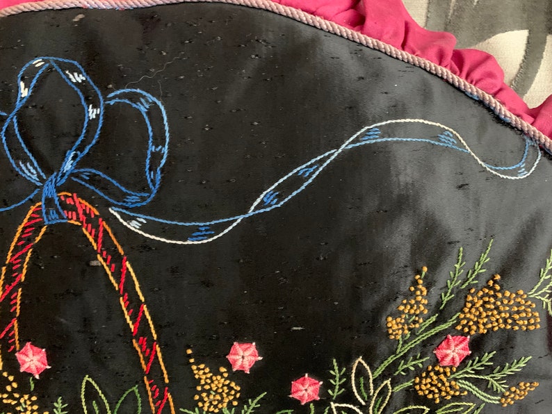 K&ouml;nnte beinhalten: Ein schwarzes Kissen mit gestickten Blumendesigns in den Farben Rosa, Gelb, Gr&uuml;n und Blau. Ein blaues Band ist oben auf dem Kissen zu einer Schleife gebunden.