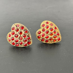Op de afbeelding: Een paar hartvormige oorbellen met rode strass-accenten. De oorbellen zijn goudkleurig en hebben een vintage stijl.