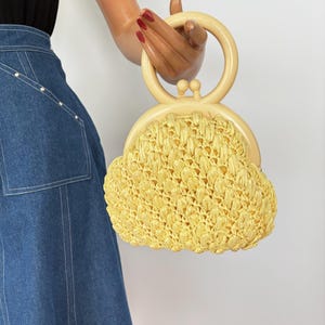 May include: A vintage yellow raffia handbag with a cream-colored handle. The bag has a rounded shape and a clasp closure. The handle is a circular design. The bag is held by a mannequin hand.