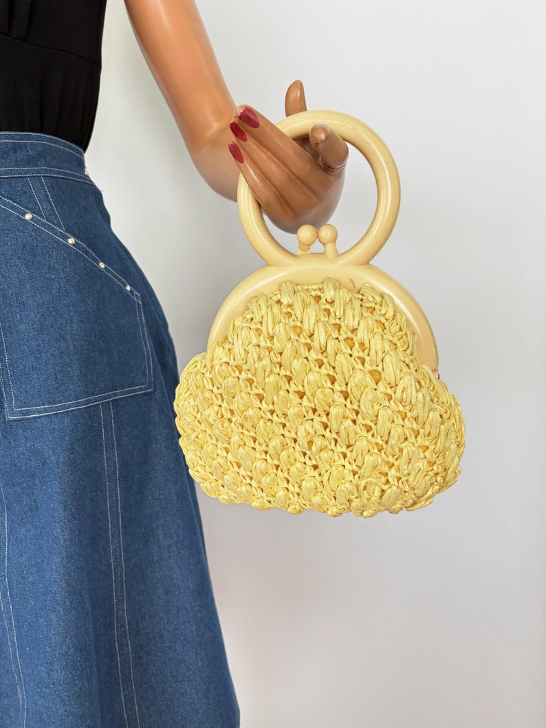 May include: A vintage yellow raffia handbag with a cream-colored handle. The bag has a rounded shape and a clasp closure. The handle is a circular design. The bag is held by a mannequin hand.