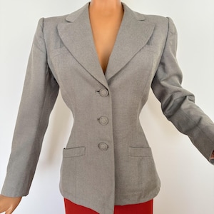 Puede incluir: Una blazer gris con un corte entallado, con solapa de muesca, cierre de tres botones y dos bolsillos delanteros. La blazer tiene mangas largas y una silueta estructurada. La blazer se combina con una falda roja.