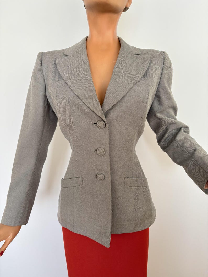 Puede incluir: Una blazer gris con un corte entallado, con solapa de muesca, cierre de tres botones y dos bolsillos delanteros. La blazer tiene mangas largas y una silueta estructurada. La blazer se combina con una falda roja.